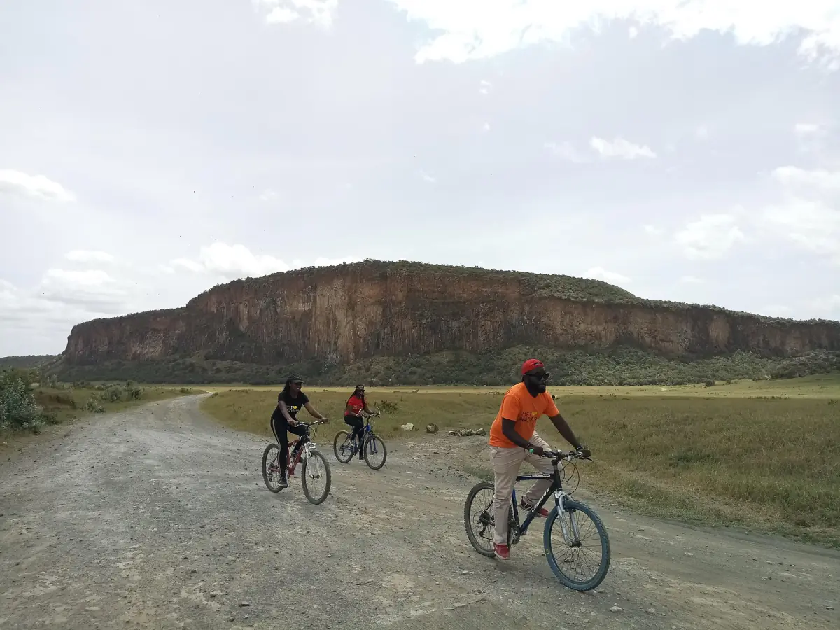 Three cyclists riding a dirt track toward a flat-topped cliff in Hell's Gate