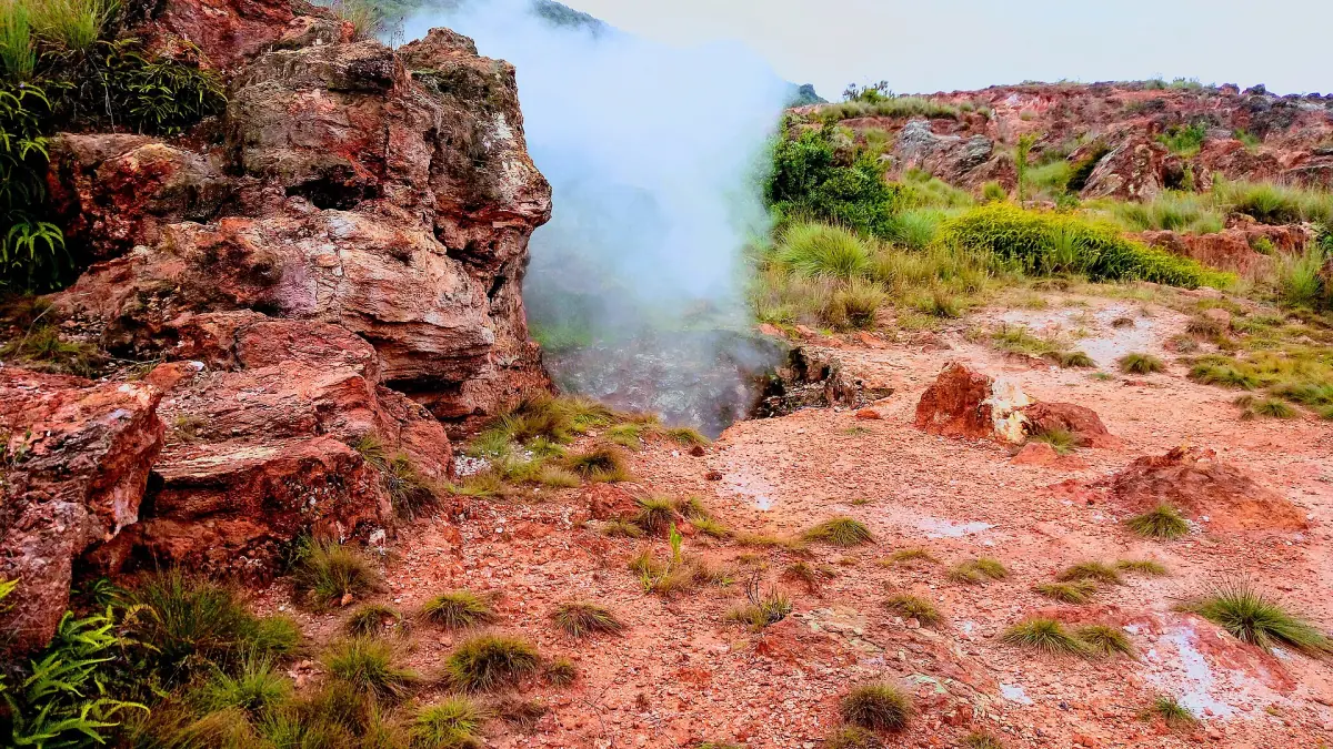 Steam rising from a vent among red volcanic rocks and green grass